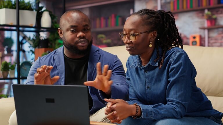 Black young couple attending an online video call connection at home, sitting on the couch and talking to their friends. Man and woman waving on a video conference, lifestyle. Camera B.